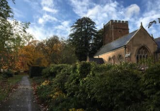 A beautiful church sits half hidden by a tumbling hedgerow. Behind it are autumnal trees with yellow leaves