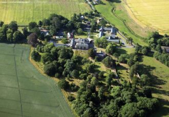 A view of a large stately home and grounds from above with fields and trees.