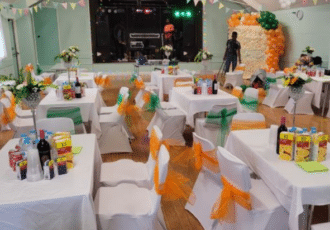 Several tables & chairs decorated with white, green & orange fill a hall. A band sets up lights and speakers on a stage.