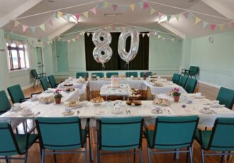 Tables laid out in a hall for afternoon tea. Large silver balloons read '80'