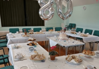 Tables laid out in a hall for afternoon tea. Large silver balloons read '80'
