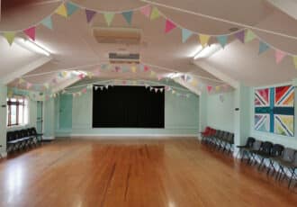 A wide, bright hall painted pale green. Brightly coloured pastel bunting hangs from the ceiling. There is a stage with black curtains on the back wall.