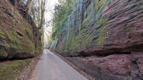 A road runs between two roughly hewn walls of red stone, moss and ivy grow down them