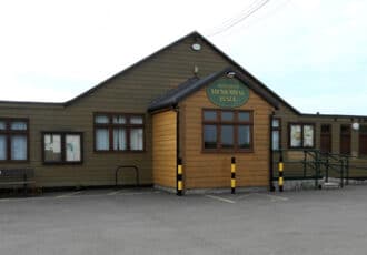 A wooden clad building with a pointed gable roof. Two green signs read "Memorial Hall" and "Nynehead Club"