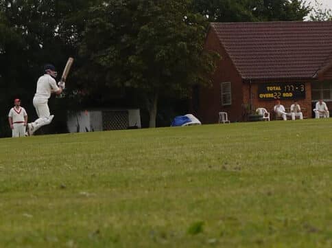 A cricket player in full outfit runs forward with his bat ready to hit the ball. Other players sit and watch by the brick club house.