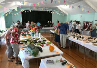 A large hall full of tables covered in fruit, vegetables, flowers and foods to be judged. People crowd around to look at it all.