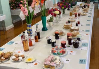 A long white table with a large selection of home made cakes, preserves & drinks. Tall, colourful gladioli stems fill the left hand side.