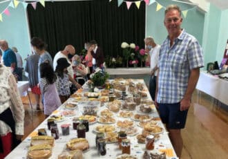 A man in a blue checked shirt smiles at the camera, he is standing in front of a table full of cakes, biscuits and scones that are ready for judging.