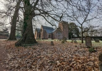 A beautiful red brick church sits in a churchyard full of fallen leaves. A bare tree fills the front of the frame. The light is wintery and grey.
