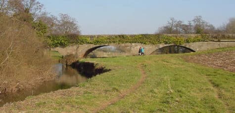 An old stone bridge lies beyond a grassy field, three arches cross the river,