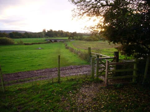A kissing gate leading to a footpath, with a view of Three Arch Bridge