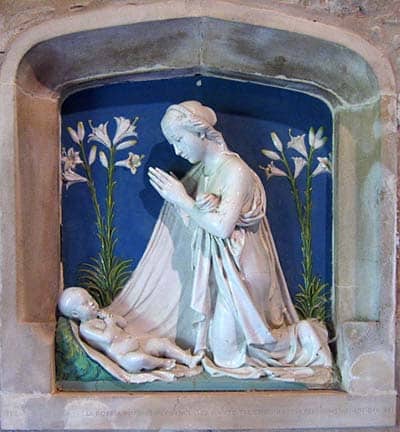 A ceramic sculpture in white on a blue background with flowers. Mary prays over the infant Jesus, swaddled in her cloak.