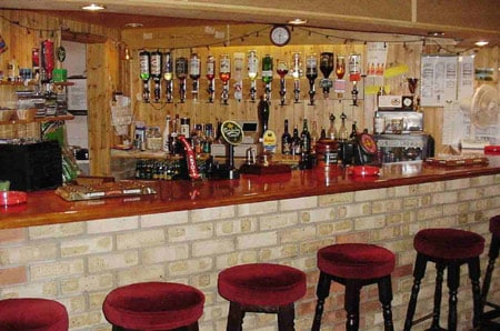 A white brick bar with a glossy wooden top, several red velvet bar stools are lined up in front, and you can see many bottles of spirits on the back wall.