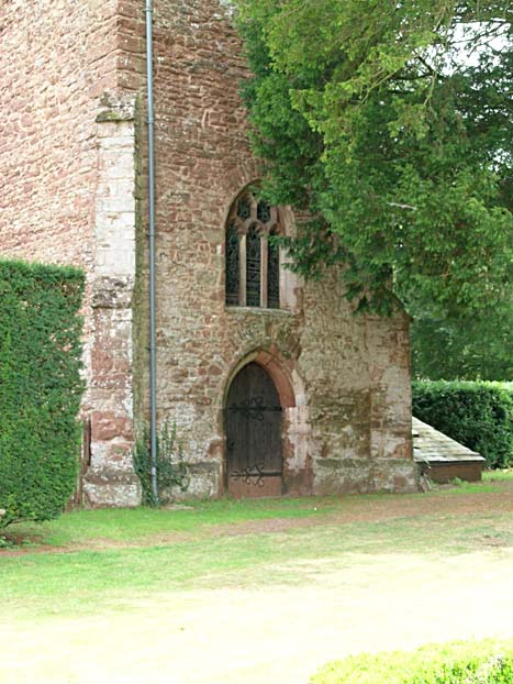 A high red brick wall, with a stained glass window and arched wooden doorway.